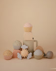 Mushie Suction Spinner Toys, shaped like rainbows, clouds, and a sun, are arranged on a beige background. Their pastel colors and food-grade silicone design create a calm, playful sensory experience.