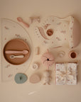 Flat lay of mushie's Silicone Placemat in pastel tones, shown with a bib, sippy cup, snack container, pacifier, teether, toys, and a floral-patterned cloth—all neatly arranged on a beige background.