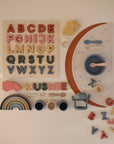A pastel flat lay features mushie Silicone Placemats topped with toddler toys—an alphabet puzzle, stacking cups, rainbow arches, toy vehicles, utensils, and loose letters—arranged on a white surface.