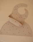 The mushie Silicone Placemat and beige baby bib showcase small black and yellow bumblebee patterns on a plain beige, food-grade silicone surface.