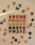 The Mushie Wooden Puzzle features colorful letters, with some placed in the board and others arranged in a circle around it on a light surface. This educational toy helps children develop fine motor skills.