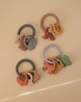 Four mushie Nature Teething Ring sets, each with animal, rainbow, and sun charms in soft pastel silicone, are arranged on a beige surface—perfect sensory baby toys for soothing little gums.
