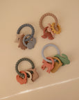 Four mushie Nature Teething Ring sets, each with animal, rainbow, and sun charms in soft pastel silicone, are arranged on a beige surface—perfect sensory baby toys for soothing little gums.