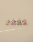 Four mushie Dog Figurine Teether toys in pastel pink, lavender, green, and peach are lined up on a cream surface, all facing away from the camera.