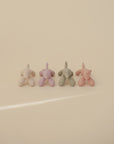 Four mushie Dog Figurine Teether toys in pastel pink, lavender, green, and peach are lined up on a cream surface, all facing away from the camera.