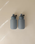 Two identical mushie Silicone Puree Pouches with matte gray looped lids sit side by side on a beige surface, resembling sleek reusable baby food pouches, softly lit with faint background shadows.