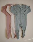 Three mushie Zipper Pajamas with feet in dusty rose, cream, and light teal are laid out side by side on a neutral background. These cozy long-sleeve onesies offer soft, comfortable sleepwear for babies.