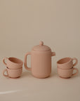The Mushie Silicone Tea Play Set features a minimalist pink design with a ribbed pattern, perfect for pretend play or a play kitchen. Set includes a teapot with lid and four stackable teacups, displayed on a beige background.