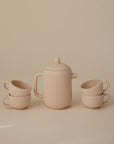 The Mushie Silicone Tea Play Set includes a beige teapot with lid and handle, surrounded by four matching ribbed cups. Perfect for play kitchens or pretend play, the set is displayed against a neutral background.