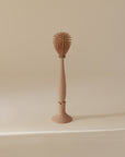 The mushie Silicone Baby Bottle Brush, featuring spiky silicone bristles and an oval head, stands upright in its matching holder against a plain beige background.