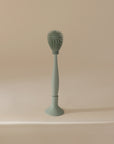 The mushie Silicone Baby Bottle Brush in sage green features silicone bristles and a spiky, rounded head, standing upright on a matching base against a beige background.