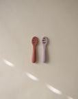 Two small Mushie First Feeding Baby Spoons, one pink and one lavender, with rounded handles and head openings, are placed side by side on a neutral background with soft light streaks.