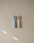 Two First Feeding Baby Spoons by Mushie, one light blue and one beige with short handles, rest side by side on a plain, light beige surface under soft lighting that casts gentle shadows.