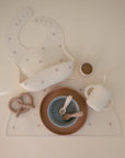 The mushie Silicone Placemat set includes a bib, plate, bowl, cup with straw, utensils, and two toy pretzels in neutral colors—all crafted from food-grade silicone and displayed on a cream-colored surface.