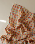 The mushie Organic Cotton Muslin Swaddle Blanket features a colorful geometric pattern of small squares and circles in orange, yellow, blue, and white on soft organic cotton, displayed on a light background with delicate folds.