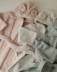 Three mushie Bear Robes in pastel pink, light gray, and sage green are displayed side by side. Each toddler bathrobe has a belt and cute bear ears on the hood for an adorable touch.