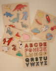 Four Mushie Wooden Puzzles—dinosaurs, space objects, farm animals, and a colorful alphabet design—are arranged on a light surface. These educational toys help develop fine motor skills.