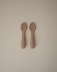 Two identical Mushie First Feeding Baby Spoons in muted brown lie side by side on a plain, light background. Each silicone teething spoon has raised sensory bumps on the bowl and "mushie" displayed on the handle.
