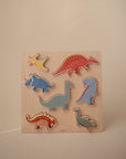 A wooden puzzle board featuring colorful dinosaur pieces, including a pterodactyl, triceratops, stegosaurus, brachiosaurus, spinosaurus, T-rex, and an aquatic dinosaur, set against a plain background.