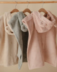 Three mushie Bear Poncho Towels in beige, sage green, and pink hang on wooden hangers. Made from organic cotton, each towel features cute bear ears for a charming touch against a neutral backdrop.