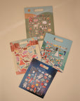 Four mushie Reusable Sticker Sets are displayed on a beige surface, each featuring colorful puffy stickers with farm animals, dinosaurs, vehicles, and space themes. The sets are arranged in a loose fan shape.