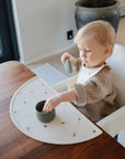 A toddler with blonde hair sits in a highchair at a wooden table, wearing a beige sweater and bib, holding a green ribbed cup with lid, and using the mushie Silicone Placemat made from food-grade silicone underneath.