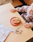 A young child wearing the mushie Long Sleeve Bib sits at a wooden table, smiling. In front of them is a pastry on a plate, while a light-colored bag with a strap rests beside them.