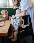 A woman in a light blue dress helps a baby in a patterned bib sit in a high chair at a wooden table. On the table, the mushie Water Resistant Wet Bag rests nearby, while a young child smiles at the baby amid food and a small vase.