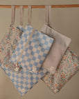 Four mushie Water Resistant Wet Bags hang from a wooden rod. The set features blue checkered, floral, and pale gold star patterns—ideal for diaper bag storage with zippered closures.