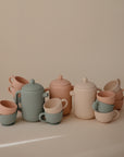 The Mushie Silicone Tea Play Set features pastel-colored cups, teapots, and mugs with ribbed textures, neatly arranged on a beige background—ideal for pretend play or a play kitchen.