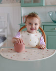 A smiling toddler with a polka-dot bib sits at the table in a pastel kitchen, holding a pink shell-shaped toy and using the mushie Silicone Placemat.