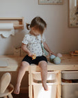 A young child with light brown hair in pigtails sits on a wooden desk holding the mushie Mix and Match Cupcake Toy, surrounded by pastel play kitchen decor and silicone cupcakes.