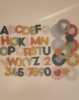 Colorful wooden letters, numbers, and pastel shapes brighten a beige wall, creating a playful display with educational fun—just like the mushie Foam Bath Puzzle offers during bath time. Decorative and engaging for kids!.