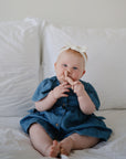 A baby in a blue dress and white headband sits on a white bed, smiling and playfully putting a finger in their nose while holding the mushie Unicorn Figurine Teether.