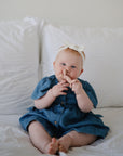 A baby in a blue dress and white headband sits on a white bed, smiling and playfully putting a finger in their nose while holding the mushie Unicorn Figurine Teether.