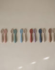 Nine pairs of Mushie First Feeding Baby Spoons in pastel colors are neatly lined up on a neutral surface, each pair showing identical self-feeding spoons with gentle lighting and soft shadows.