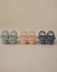 Three pairs of pastel baby shoes—light green, pale pink, and muted blue—are lined up against a neutral background, mirroring the soft hues of popsicles made with the mushie Toddler Ice Pop Tray silicone mold.