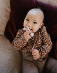 A smiling baby in a floral shirt and brown pants sits on a couch with a maroon pillow, holding the mushie Cat Figurine Teether to their mouth.