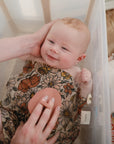 A smiling baby with reddish hair enjoys bath time wearing a floral-patterned cover, while an adult uses mushie Cradle Cap Brushes with silicone bristles on the baby's chest.