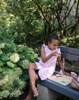 Two young children sit on a garden bench, one painting while the other watches. Nearby, a Mushie Wooden Puzzle boosts their fine motor skills, surrounded by green foliage and blooming hydrangeas.