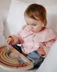 A toddler in a pink shirt and jeans sits on a white bed, engaged in screen-free play with the mushie Wooden Magnetic Rainbow Maze, a toy designed to develop fine motor skills.