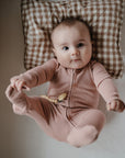 A baby in mushie Zipper Pajama lies on a checkered brown pillow, curiously holding one foot while resting on a white bed.