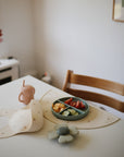 A child’s table set with a wooden chair, silicone bib, mushie Silicone Placemat, divided plate of cucumber, tomatoes, oranges, spill-proof cup, and a flower-shaped spoon rest.
