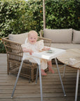 A smiling baby sits in a white high chair on a wooden patio. On the table nearby, the mushie Suction Spinner Toy offers sensory play, while a small plate and mug are set next to a wicker sofa with greenery in the background.