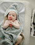 Baby on changing pad with blue floral towel, teether, and mushie Diaper Caddy nearby.