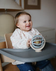 Child sitting in a high chair holding a suction spinner ball toy, smiling.