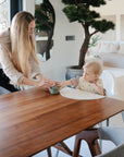 A woman with long blonde hair hands a cup to a toddler in a high chair at a wooden dining table, where mushie Silicone Placemats add style and easy cleanup to the bright, modern space filled with plants and neutral decor.