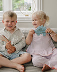 Two smiling kids on a couch: one holds colored pencils, the other a mushie Silicone Sensory Toy.