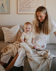 A woman in a white eyelet dress sits on a beige couch holding her smiling baby, who is wrapped in a mushie Organic Cotton Muslin Swaddle Blanket, surrounded by soft pillows, a light blanket, and framed art in the background.