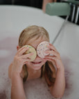 A smiling child in a bathtub holds two round bath bombs over their eyes, surrounded by bubbles—just like pretending to decorate mushie’s Mix and Match Cupcake Toy cupcakes in a play kitchen, sparking creativity and supporting fine motor development.